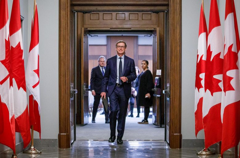 Tiff Macklem entre avec assurance dans la Chambre des communes. Des drapeaux canadiens bordent le corridor.