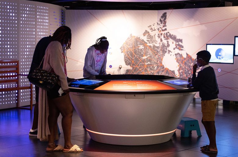Quatre personnes interagissant avec des tablettes numériques reliées à une table circulaire au Musée de la Banque du Canada.