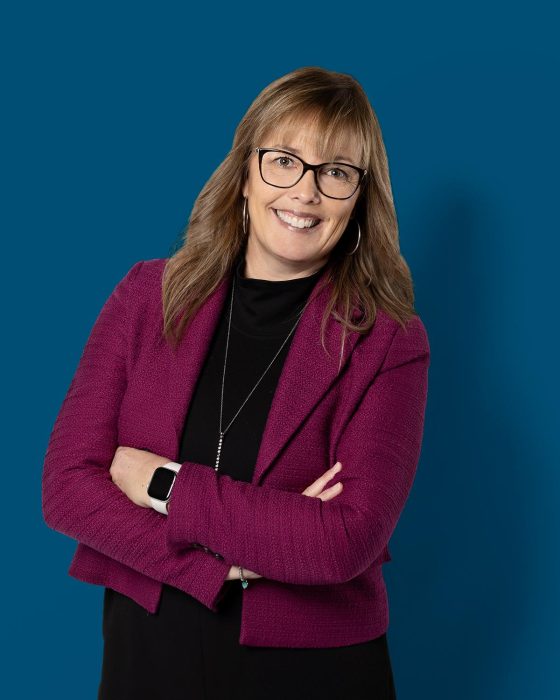 Une femme d’âge moyen aux cheveux blonds et à la peau claire, en tenue professionnelle, sourit chaleureusement à la caméra, se montrant amicale et facile d’approche.