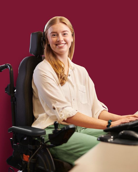 Une jeune femme installée en fauteuil roulant derrière son bureau sourit à la caméra en travaillant sur l’ordinateur.